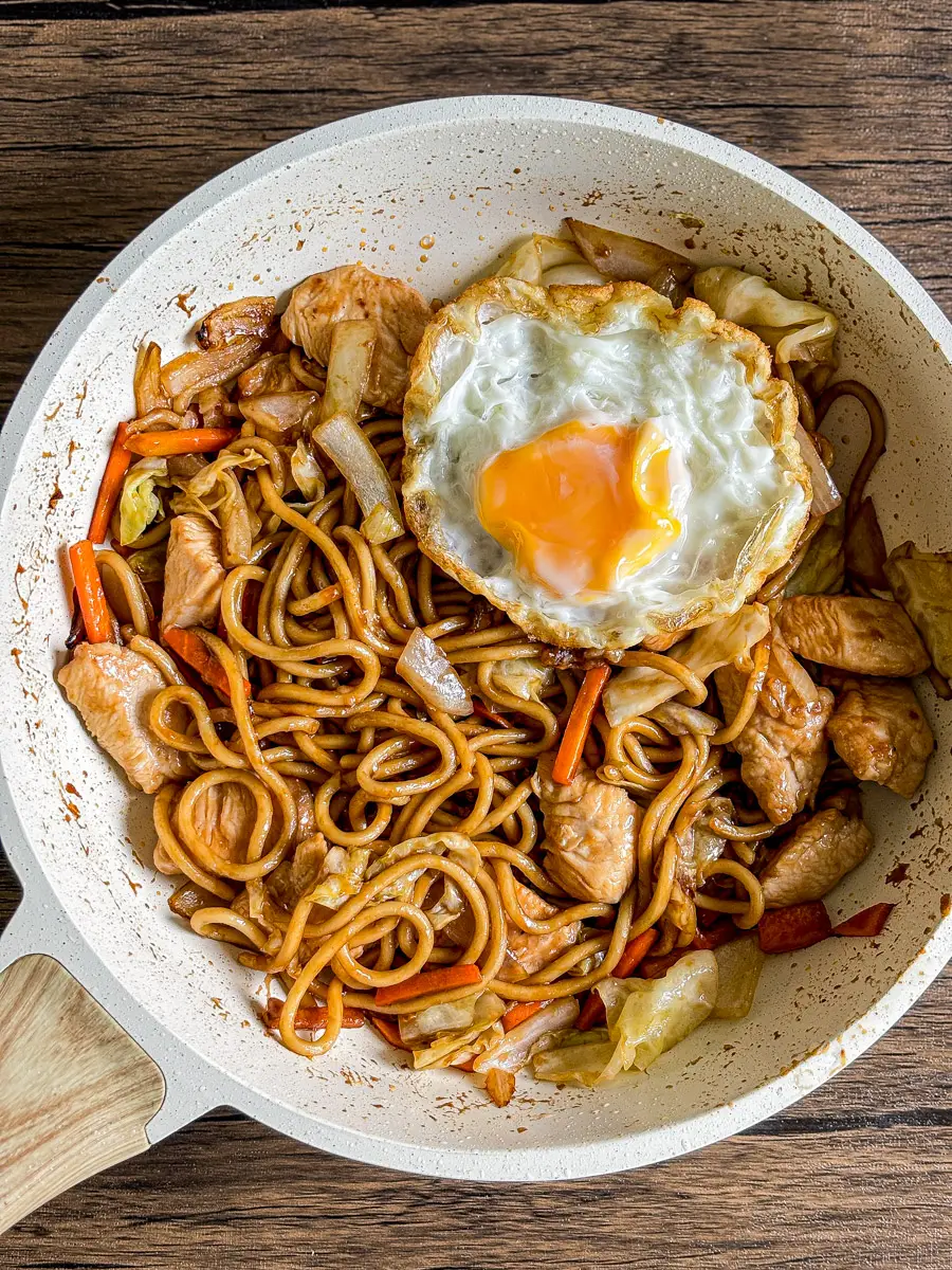 Chicken yakisoba noodles stir-fried with vegetables and topped with a crispy fried egg in a pan.