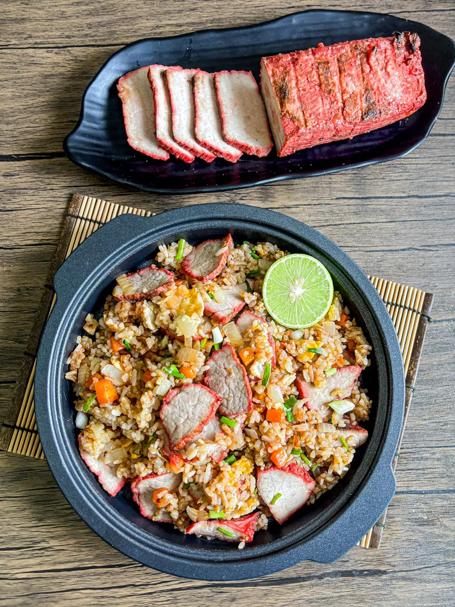 Top-down view of homemade char siu fried rice served in a black bowl with lime, next to sliced Chinese BBQ pork on a black plate.