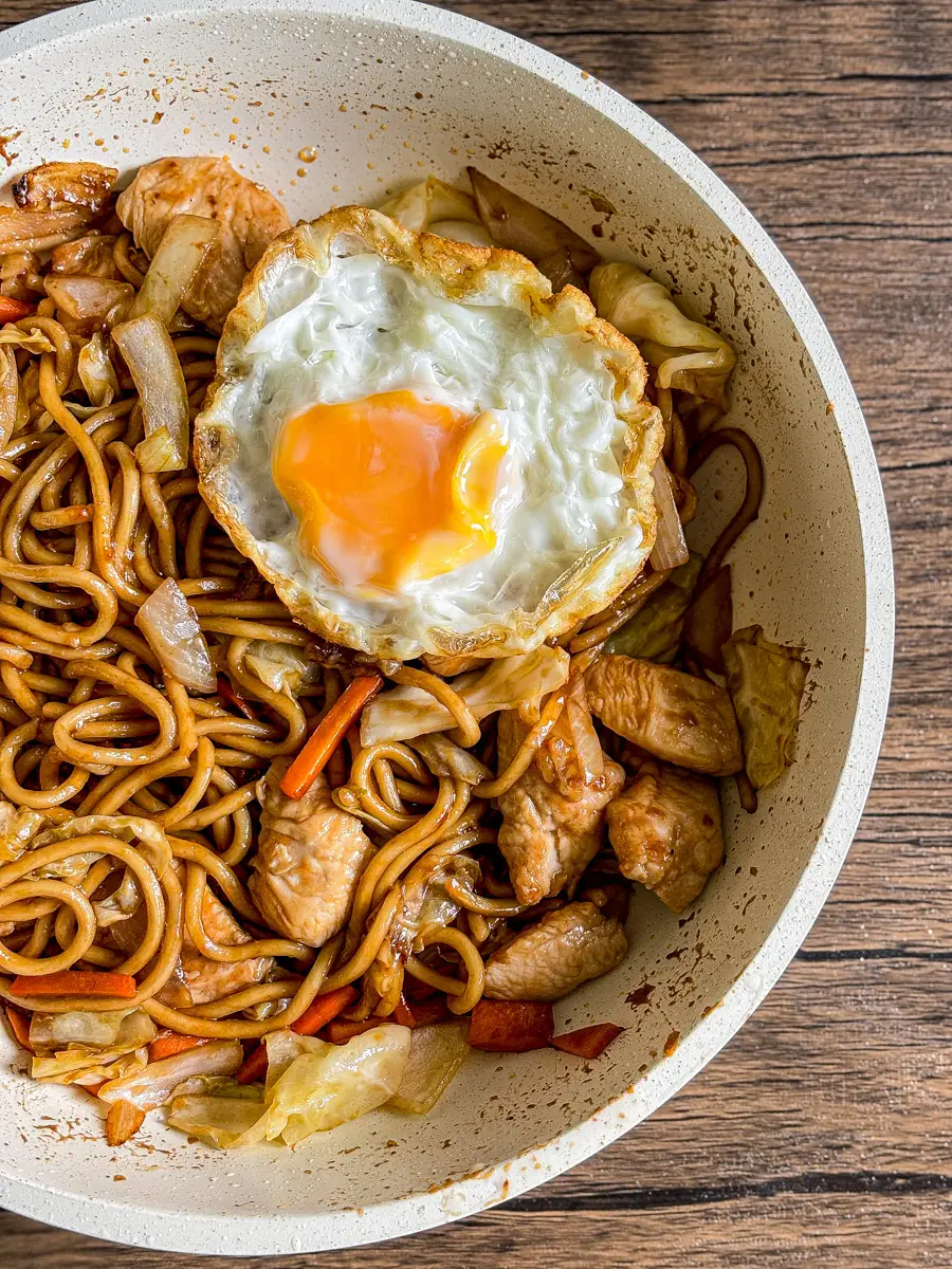 Yakisoba chicken stir-fried with vegetables and topped with a crispy fried egg in a pan.